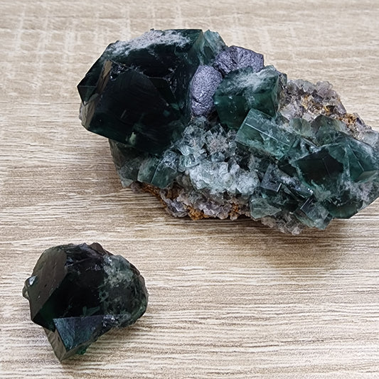 Two chunks of green fluorite crystals from The Crystalary's LIVE- CheerBow- 8/16/2024 collection are displayed on a wooden surface. The larger piece features dark green cubic crystals embedded in a matrix, while the smaller piece consists of a single, dark green cubic crystal. Resembling pocket-sized Northern Lights minis, the crystals have a semi-transparent appearance.