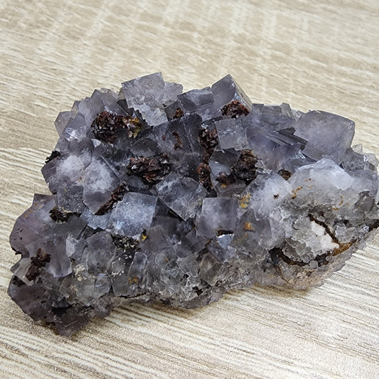 A close-up photograph of a rock specimen features numerous translucent cubic purple crystals, likely fluorite, embedded in a rough, gray matrix on a wooden surface. The crystals have a subtle sparkle under the light, capturing the essence of The Crystalary's LIVE- cherylwinsomlosesome- 6/20/24 collection.