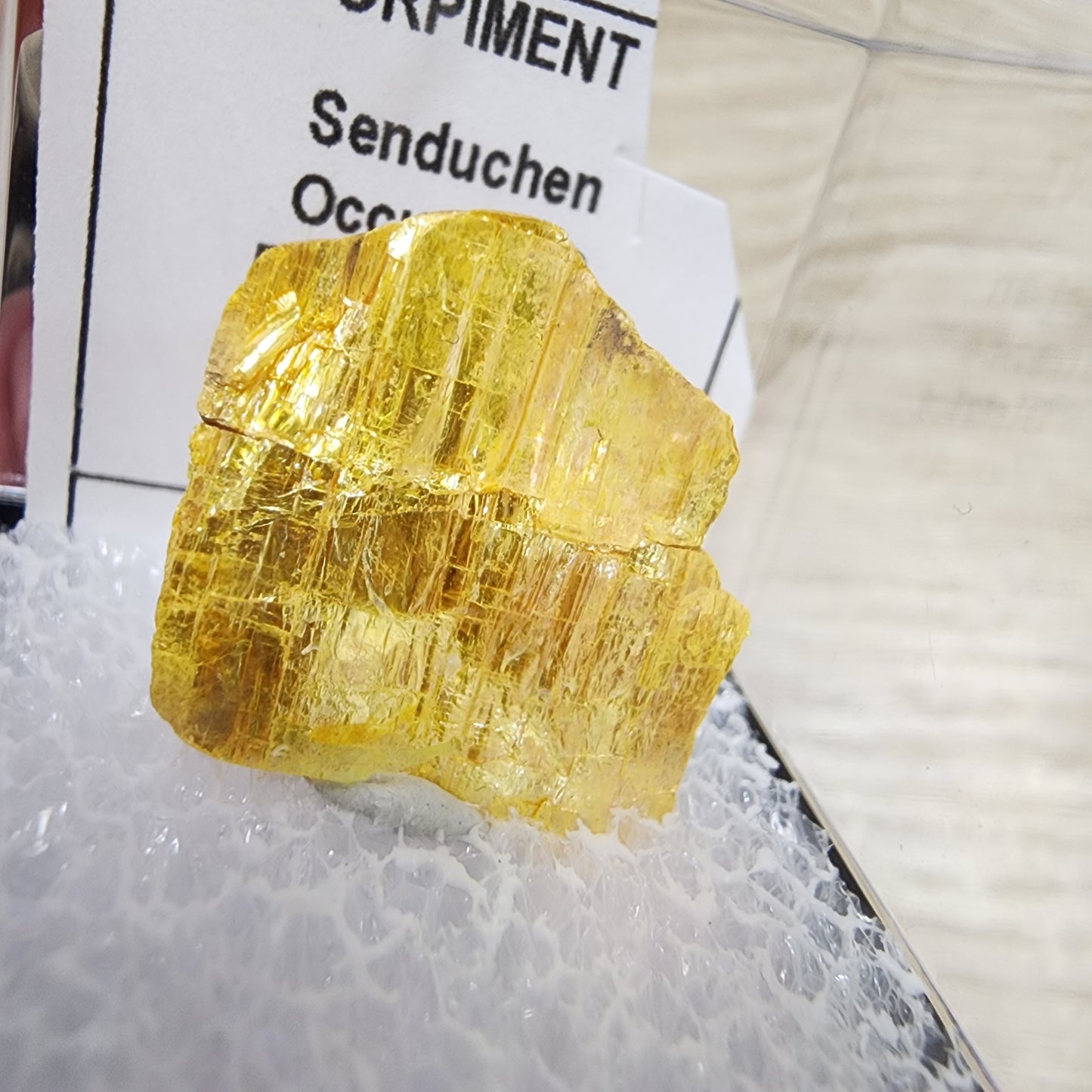 A vivid yellow orpiment mineral specimen from Senduchen, showcased atop white fluff in a compact glass or plastic case, features a crystalline texture with noticeable striations and rests on a bed of white fibrous material similar to millerite's intricate formations. Available under the brand "The Crystalary" as part of the LIVE- crystalcollectorliz collection dated 8/29/2024.