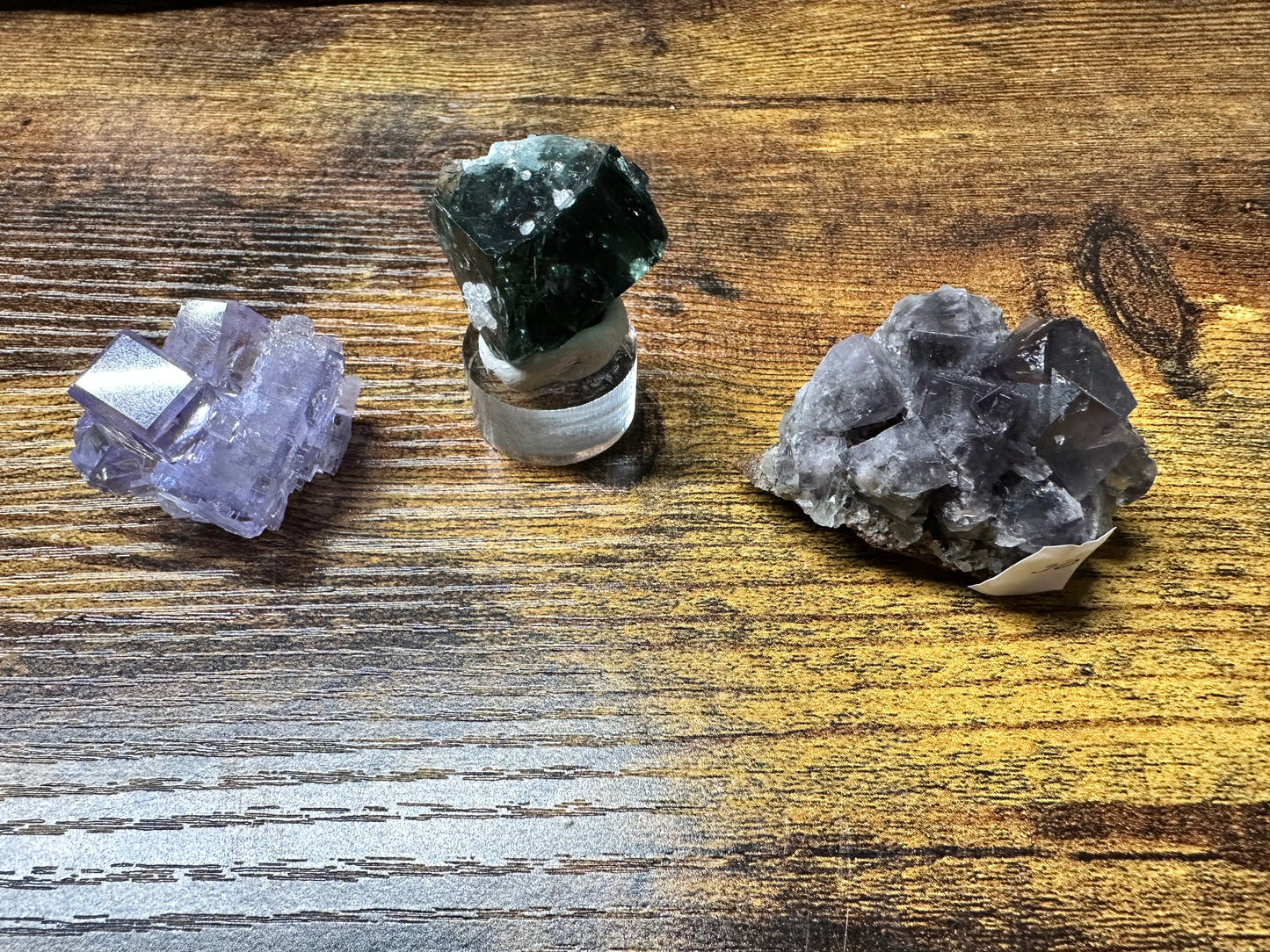 Three mineral specimens are showcased on a wooden surface. On the left is a light purple cubic crystal of fluorite. In the center is a dark green crystal sourced from Jaimina Mine in Spain, mounted on a clear stand. On the right, clusters of translucent violet crystals complete the display, all part of The Crystalary's LIVE- DocManny- 4/12/24 collection.