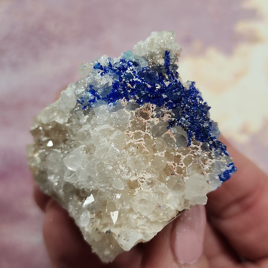 A close-up of a person's hand holding a crystalline mineral specimen from The Crystalary's LIVE collection, specifically the Eric.Jazzar models available on 1/4/24 and 12/28/23. The light-colored mineral features predominantly clear and white crystals with a striking section of vibrant blue Linarite crystals on its upper part. The background is softly out-of-focus in pink and yellow, reminiscent of the hues found in Socorro, New Mexico.