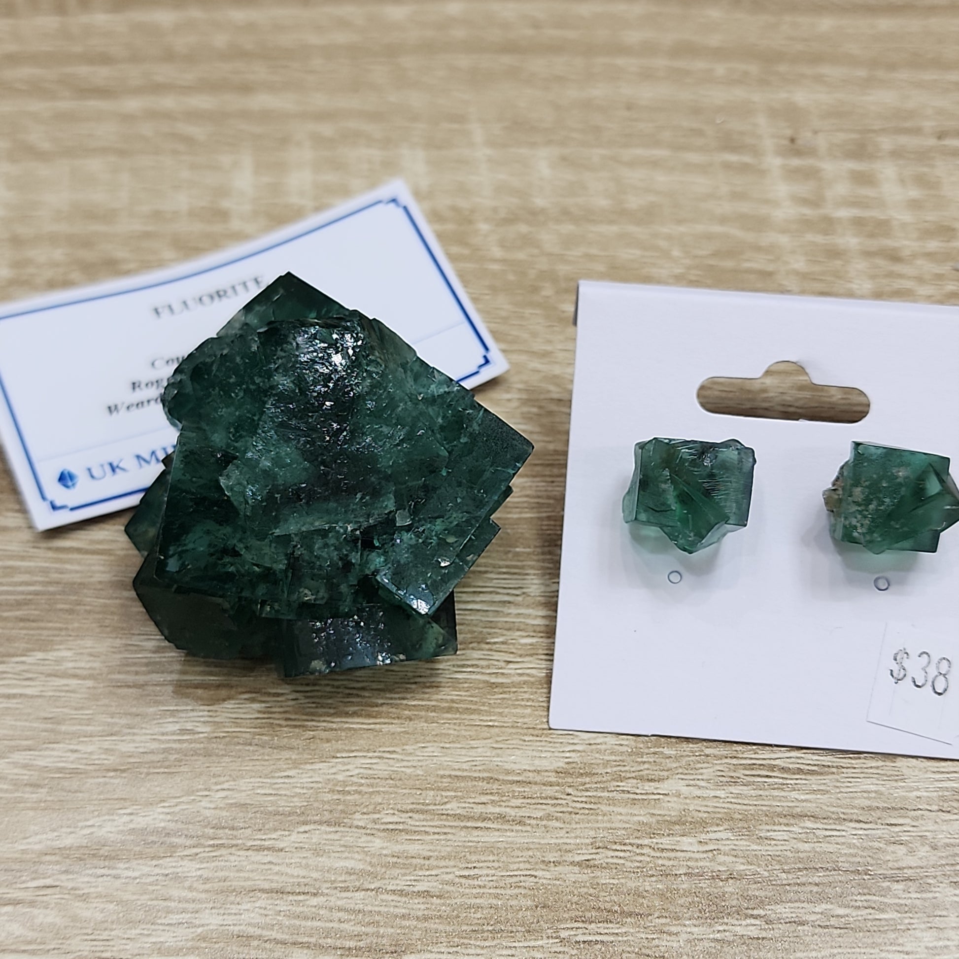 Image of The Crystalary green fluorite crystal cluster displayed on a wooden surface, with a small blue and white card behind it. Next to the crystal is a pair of The Crystalary green fluorite stud earrings on a white display card marked with a price tag of $38. Perfect for adding to your Cousin Jack Pocket collection.