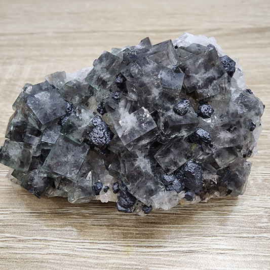 Here is a rewritten version of the sentence using the given product data:

The Crystalary's LIVE- exbeertz- 8/16/204 features a captivating cluster of clear, cubic fluorite crystals intertwined with dark, metallic galena crystals. The fluorite showcases a subtle greenish hue that evokes the beauty of the Northern Lights and is set against a rustic wooden surface. The overall texture is rough and varied, highlighting the unique character of each crystal type.