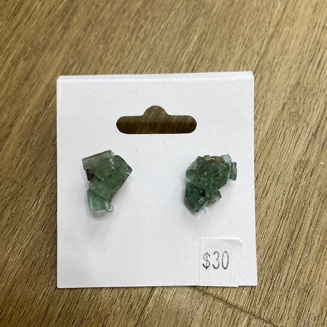 A pair of rough, green fluorite fish mineral earrings from The Crystalary's "LIVE- Fluorite Fish- 5/30/24" collection is displayed on a white card with a hole for hanging. The card rests on a wooden surface and features a small price tag in the bottom right corner indicating $30.