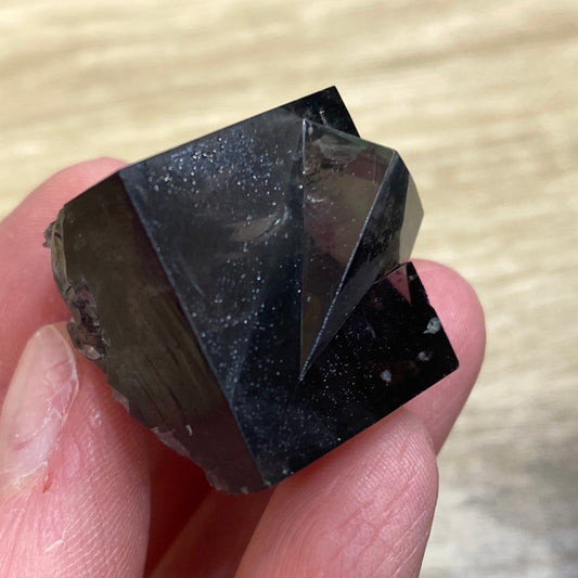 Close-up of fingers holding a dark-colored, multifaceted crystal from The Crystalary, named LIVE- It’s Just Juice- 6/6/24. The crystal boasts a shiny surface with various geometric faces, casting reflections and subtle speckles. The background is a light-colored wooden surface with the faint engraving "675" barely visible in the corner.