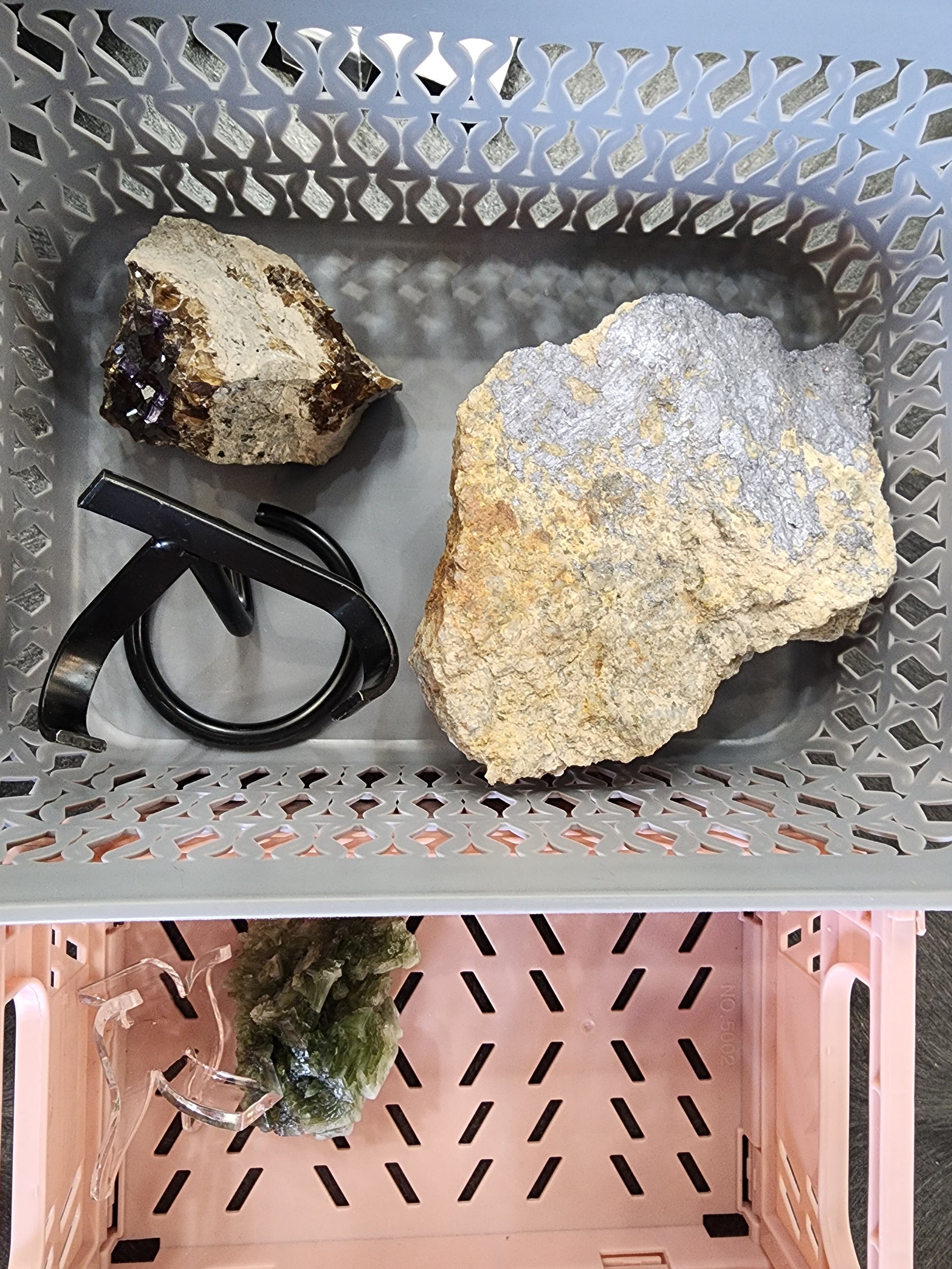 Two bins containing various objects. The top bin holds a rock with amethyst crystals, a larger rough-surfaced rock, and a pair of black-framed glasses. The bottom bin features a green, crystal-like object, possibly molybdenite, and what appears to be a clear stand. These items are part of the "LIVE- it'sjustjuice- 7/16/24" collection by The Crystalary.