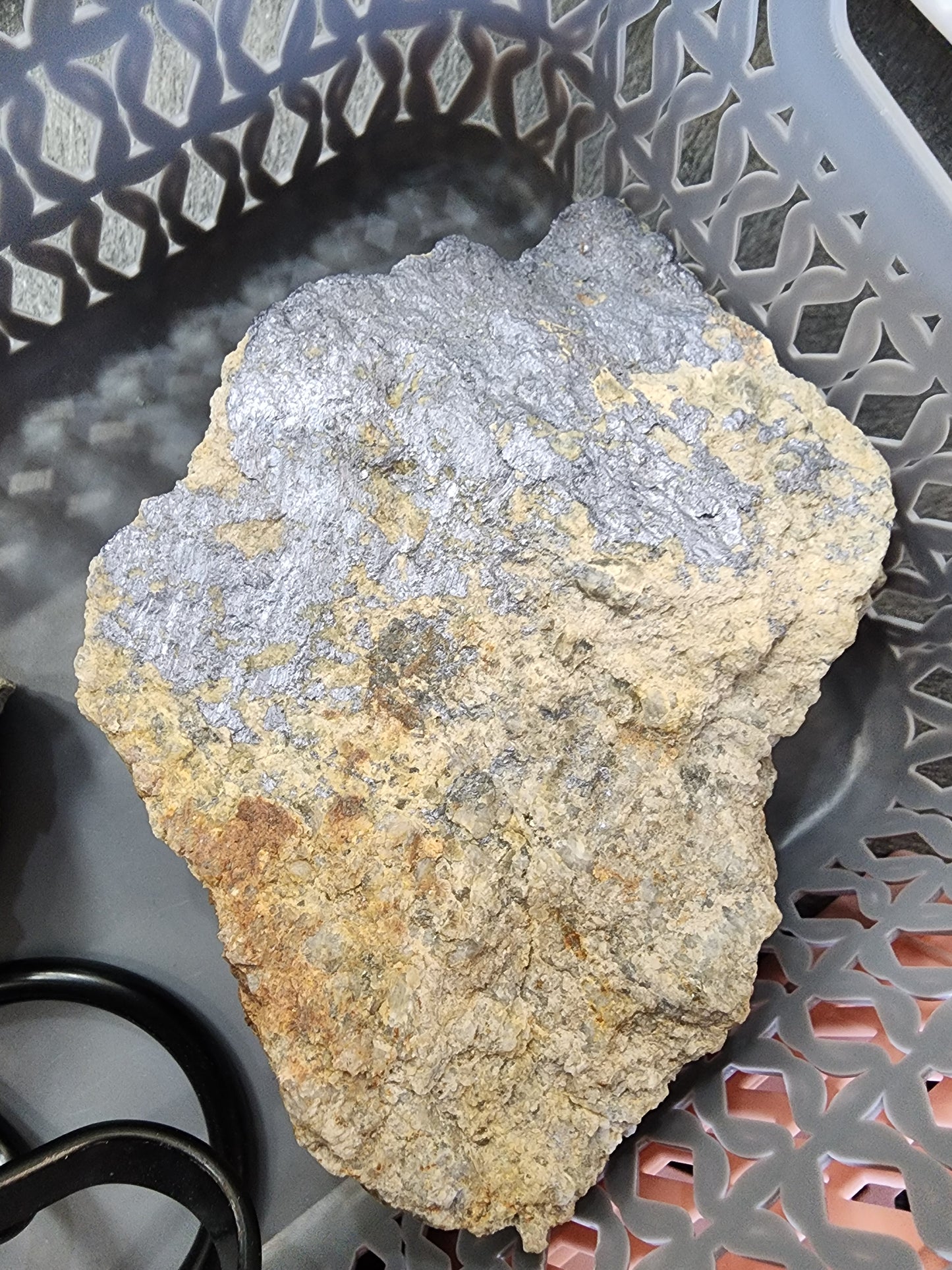 A large, rough piece of rock with a mix of gray and brownish-yellow colors, possibly containing molybdenite, sits in a perforated plastic basket labeled "LIVE- it'sjustjuice- 7/16/24" from The Crystalary. A black cable or wire can be seen partially in the corner of the image.