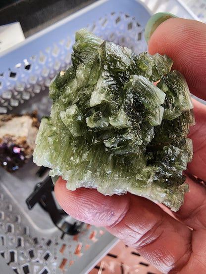 A person holding a LIVE - it'sjustjuice - 7/16/24 mineral cluster from The Crystalary, showcasing multiple pointed crystals with vibrant green color and translucent qualities similar to halite. In the background, there is a gray perforated tray featuring additional minerals or rocks.