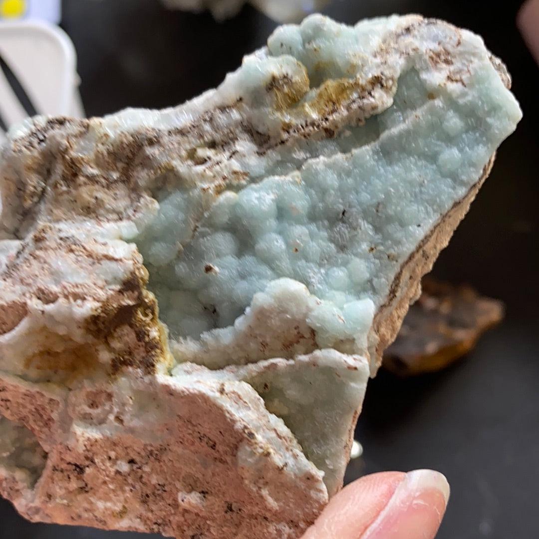 A close-up view of a person's hand holding a rock specimen from The Crystalary's LIVE-janisjoplin96boo-Oct 27 collection, showcasing calcite with light blue and white mineral formations interspersed with rough brown and gray patches. The intricate textures and varying colors stand out against the blurred background.