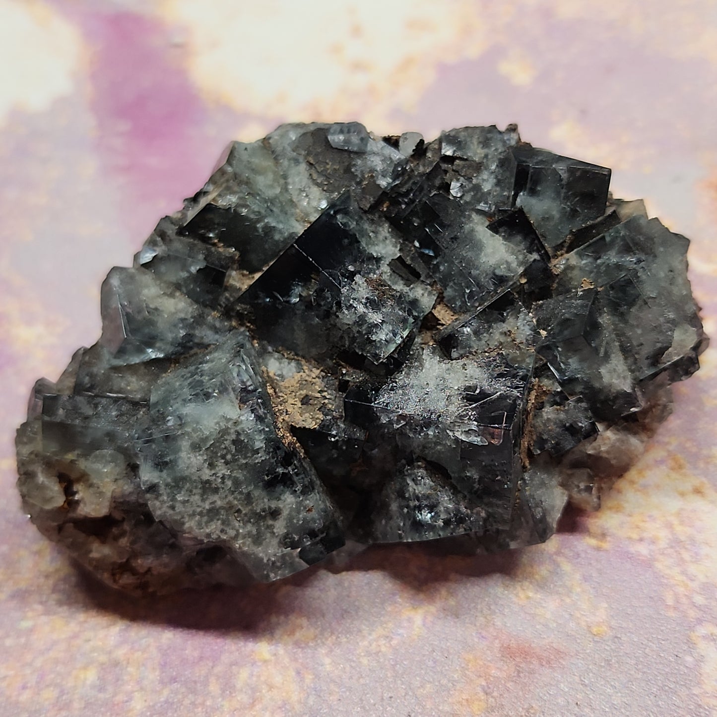 Close-up of a jagged, dark-colored mineral specimen from The Fairy Holes Pocket in The Crystalary's collection, LIVE- K(pssrevoked)- 12/26/23 paid 300. The mineral exhibits a rough, crystalline structure with sharp edges in a stunning mix of black and gray hues. The background is softly blurred with shades of light pink and beige.