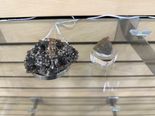 Two mineral specimens from The Crystalary's LIVE- Karen- 5/11/24 collection are displayed on transparent pedestals on a glass shelf. The left specimen is a rough, dark-colored rock with crystalline structures, likely sourced from the Smoky Rabbit Pocket, while the right specimen appears to be a polished, multi-colored fluorite stone. Both are under soft lighting.