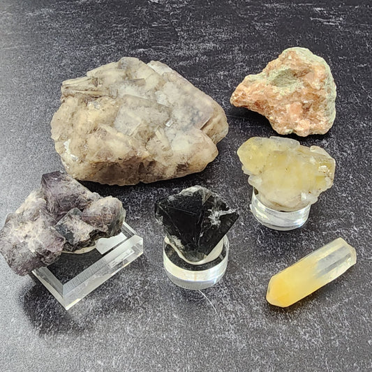 Various mineral specimens from The Crystalary, including a cubic cluster of light brown crystals, a rough orange stone, a cluster of pale yellow crystals, Mango quartz, a dark black crystal, a Purple Haze crystalline structure, and a yellow polished crystal wand are arranged on a dark surface as part of the LIVE- Karenwebdellrogala- 6/13/24 collection.