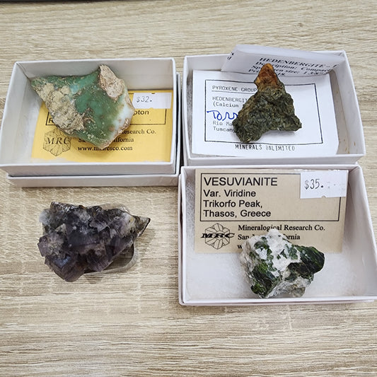 Four minerals displayed in two rows. Top row: a green vesuvianite in a thin cardboard box with a price tag of $32, and a brownish mineral in a similar box with a price tag of $35. Bottom row: a dark purple fluorite and a green-and-white mineral, both housed in thin cardboard boxes. This collection is part of The Crystalary's LIVE- karenwebdellrogala- 8/23/24 lineup.