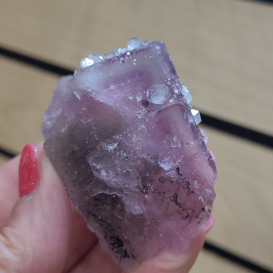 A hand with red-painted nails holds the rough, translucent purple crystal of The Crystalary's LIVE- kricketlee- 5/14/24 fluorite, its small, sparkling facets catching the light. The wooden plank background is blurred.