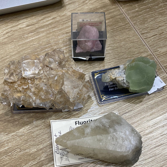 The image features several mineral specimens meticulously arranged on a wooden table. Among them are a quartz cluster, a green octahedron fluorite, a pink rhodochrosite, and a white translucent crystal, each accompanied by identifying labels. A note card labeled "Fluorite" is partially visible next to the white crystal. All these exquisite specimens are part of The Crystalary's collection showcased in their LIVE- KrickettLee- 6/6/24 display.