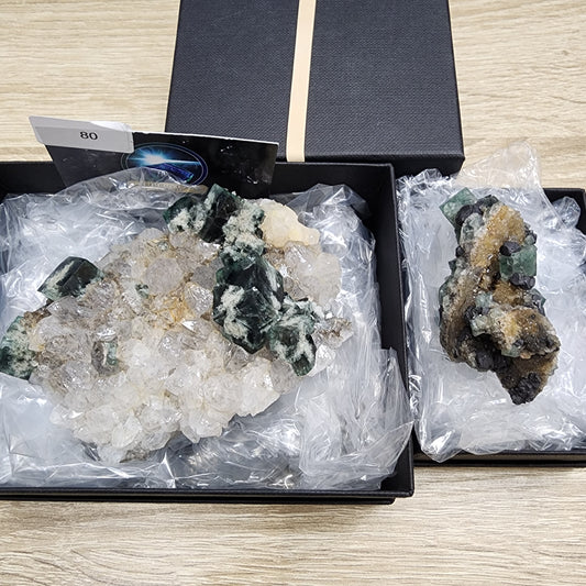 Two mineral specimens from The Crystalary's "LIVE- Lena_Crystals- 8/16/2024" collection are displayed in black boxes lined with clear plastic wrap. The specimen on the left boasts large, clear, and green crystals resembling a supernova, while the one on the right contains a blend of green and tan crystals. Both boxes are positioned on a wooden surface.