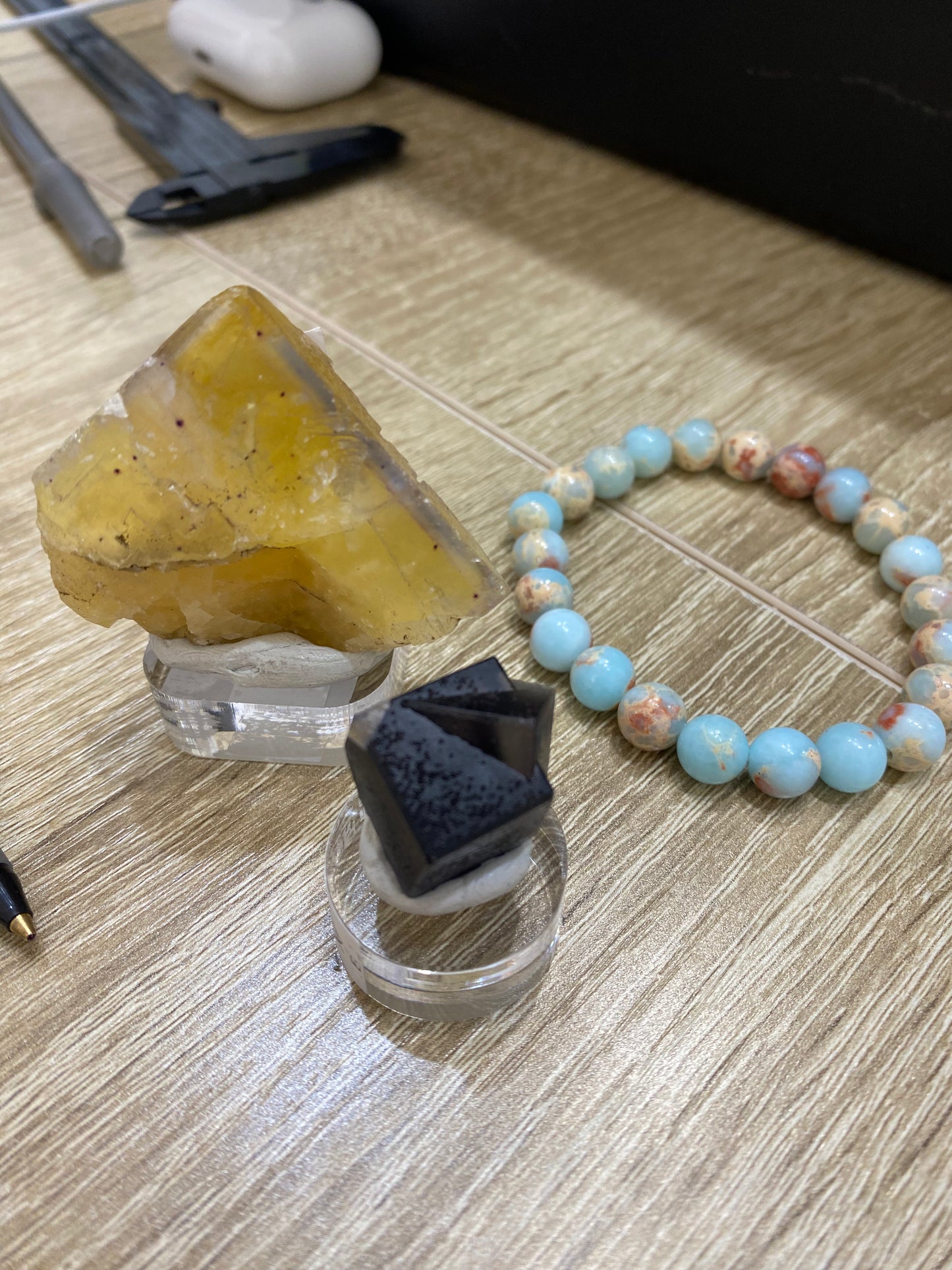 A close-up of a desk features two crystal specimens from The Crystalary's LIVE- Mandy Jo collection, dated 7/11/24. One crystal is yellow and the other black, both elegantly displayed on clear stands. To the right, a striking beaded bracelet with light blue and pinkish-white beads catches the eye, reminiscent of Shoushan Jade. Various desk accessories like a pen and earphones are visible in the background.