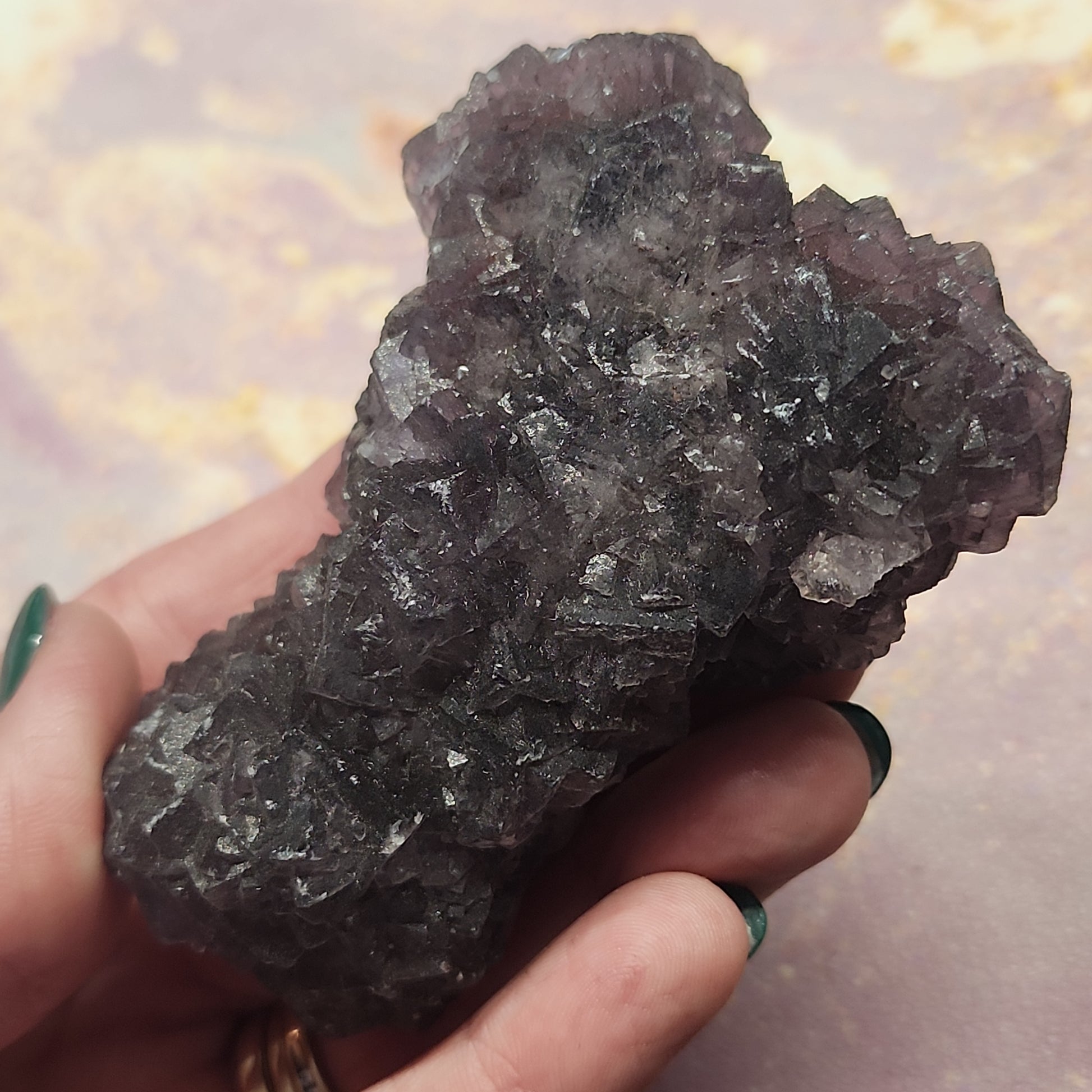 A hand with teal-painted nails holds a large, dark, and jagged mineral rock with a purplish-black hue, resembling the LIVE- melissa_mn- 12/21/23 from The Crystalary. The background is a soft, blurred mix of pastel pink and yellow colors.