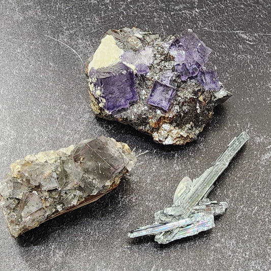 Three mineral specimens are displayed on a dark surface. One, called Purple Haze, features purple cubic crystals. Another has translucent, rough formations from Elmwood. The third is a specimen of Stibnite from The Crystalary’s LIVE- Mindy-Inked collection dated 6/13/24, showcasing metallic, slender, bladed structures with an iridescent sheen.