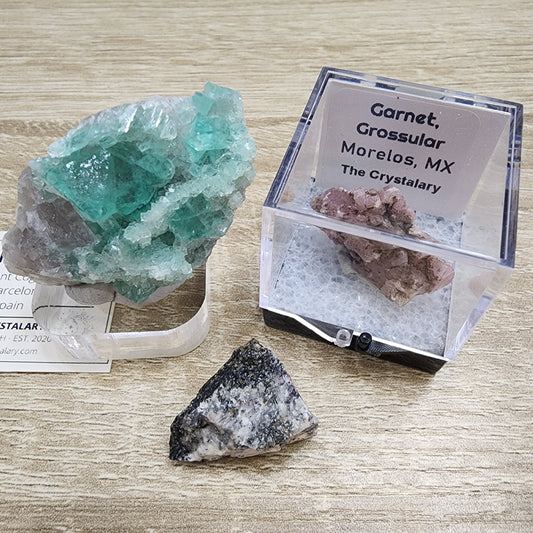 Three mineral specimens are displayed on a wooden surface. The leftmost is a large green crystal cluster. The center shows a smaller green and white rock fragment. The rightmost item is a pink Garnet labeled "Garnet, Grossular, Morelos, MX" in a clear plastic box from "LIVE- MindyInked-" by The Crystalary.