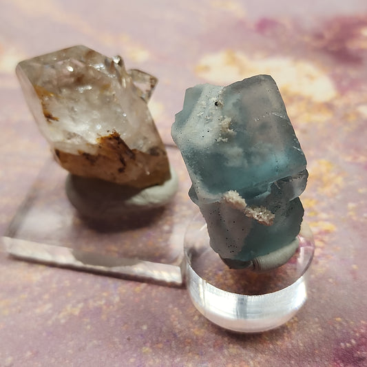 Two crystals from The Crystalary's LIVE- miriam- 12/14/23 collection are displayed on a pinkish surface. The crystal on the left is an amber-colored gem with inclusions, mounted on a square base. On the right is a blue-green crystal with inclusions, perfect for an XHL Blue giveaway, mounted on a round base.