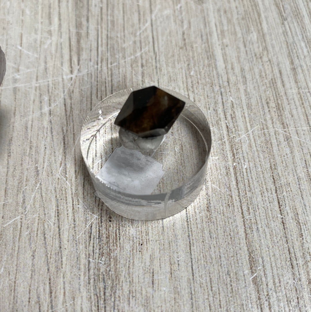 A cylindrical metal ring with a hexagonal prism-shaped Morion Quartz partially embedded in a white substance sits on a textured wooden surface. The industrial yet rustic composition contrasts the metallic and natural elements against the rough backdrop, evoking the raw beauty of Cave in Rock formations.

