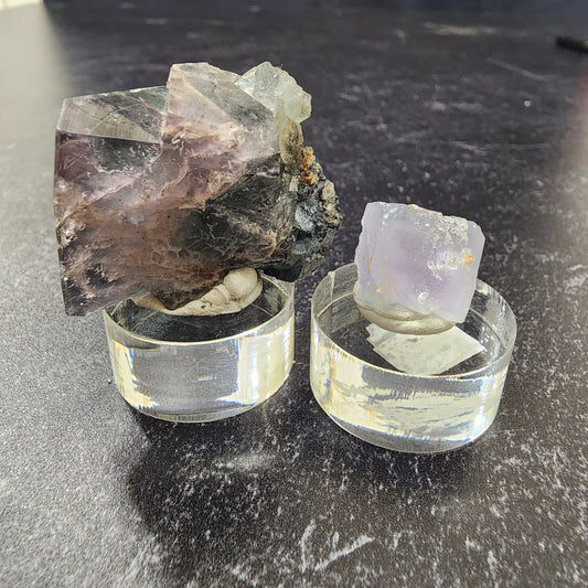 Two mineral specimens from The Crystalary's LIVE-mirrormasked-6/13/24 collection are elegantly showcased on clear display stands against a dark background. The larger specimen on the left features a dark, mottled appearance with a purple haze of black hues and boasts a rough texture. In contrast, the smaller specimen on the right is translucent, light purple, and exhibits a more uniform texture—a true pocket treasure.