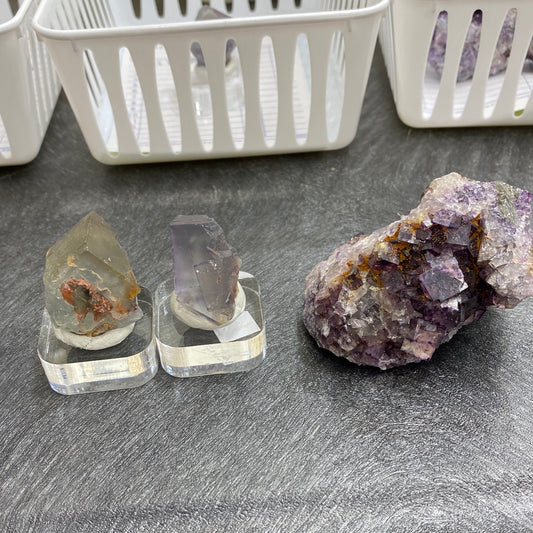 Three crystals from The Crystalary are displayed on a gray surface. Two smaller Fluorite crystals, transitioning from clear to purple hues, are elegantly placed on square stands next to a larger, multifaceted and rough purple crystal. White baskets in the background create an organized display area reminiscent of an El Hammam market, showcasing items labeled under the LIVE- missjess88- 5/11/24 collection.