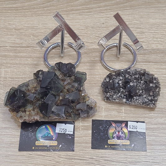 Two unique mineral specimens are displayed on a wooden surface. The one on the left, known as LIVEnor- 5/11/24 by The Crystalary, has dark, pyramid-shaped crystals with a price tag of $1250. The right specimen features smaller, clustered crystals from the Rainbow's End Pocket and is priced at $350. Each has a business card with The Crystalary logo below.