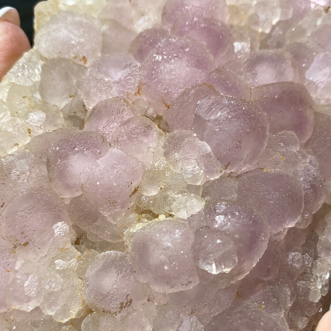 A close-up photo features a cluster of botryoidal formations of grape agate from Shangbao, showcased in the LIVE- roxonboard- 2/22/24 collection by The Crystalary. The minerals exhibit a light purple and translucent appearance, with a bumpy, rounded texture reminiscent of grape clusters. The agate has a frosted surface accented with quartz-like crystals scattered throughout.