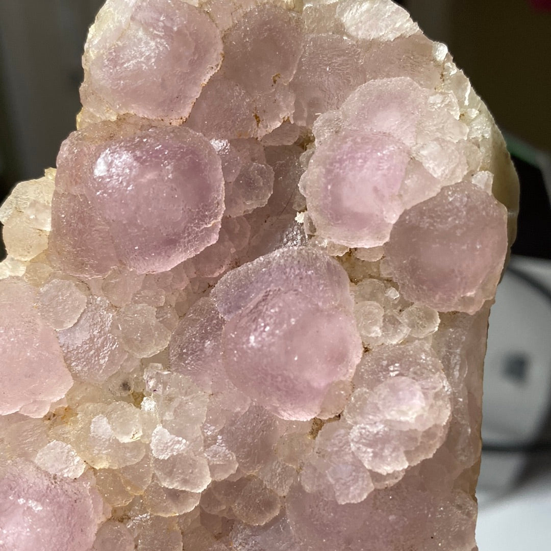 Close-up image of a LIVE- roxonboard- 2/22/24 mineral specimen by The Crystalary, showcasing grape-like clusters of translucent, light pink FLUORITE formations on a crystalline backdrop. The formations appear rounded and smooth, creating a visually intriguing contrast with the rugged crystal surface.