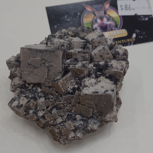 A cluster of cubic mineral crystals with a dark gray and white speckled appearance is displayed on a flat surface. In the background, there is a piece of paper or card with some text, an image of the LIVE- Sammio- 5/10/24 from The Crystalary, and a price tag showing $86.00.