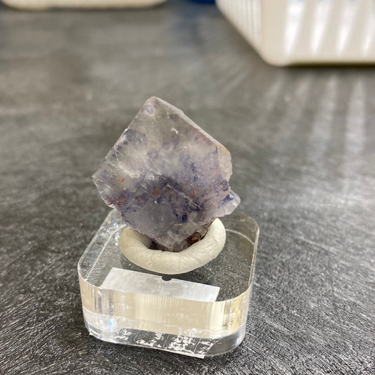 A mineral sample labeled "LIVE- Sammio- 5/11/24" from The Crystalary, possibly sourced from El Hammam, is elegantly showcased on a small, clear stand. Weighing 32 grams, the roughly pyramid-shaped crystal boasts a smooth surface and displays a transition from transparent to translucent with accents of purple and gray. The textured gray background adds an exquisite touch to its presentation.