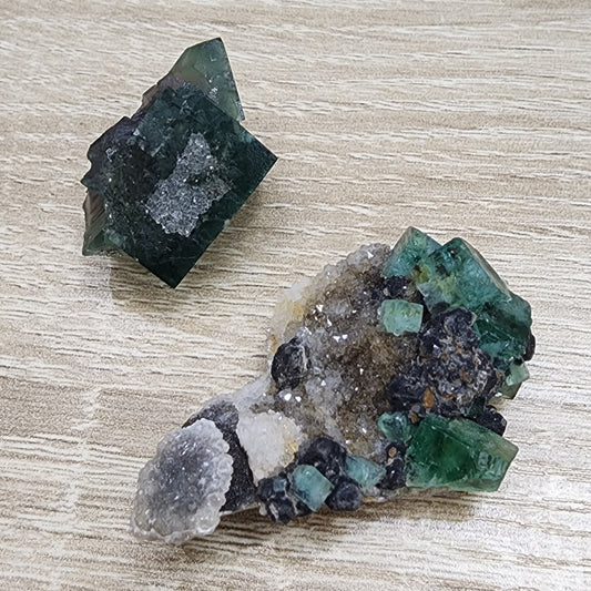 Displayed on a wooden surface are two mineral specimens from The Crystalary's LIVE-sinfullilblonde-8/16/2024 collection. The top left specimen, reminiscent of mini-20 Northern Lights, showcases green cubic crystals, while the bottom right specimen features a diverse array of clear, white, and green crystals in various shapes and sizes.