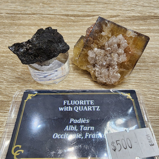 Two mineral specimens from The Crystalary rest on a wooden surface. According to the labels, one is a spectacular fluorite with quartz from Padies, Albi, Tarn, Occitanie, France. Priced at $500 and listed as LIVE- sinfullilblonde- 8/23/24, one specimen appears dark and rough while the other is lighter with visible quartz crystals.