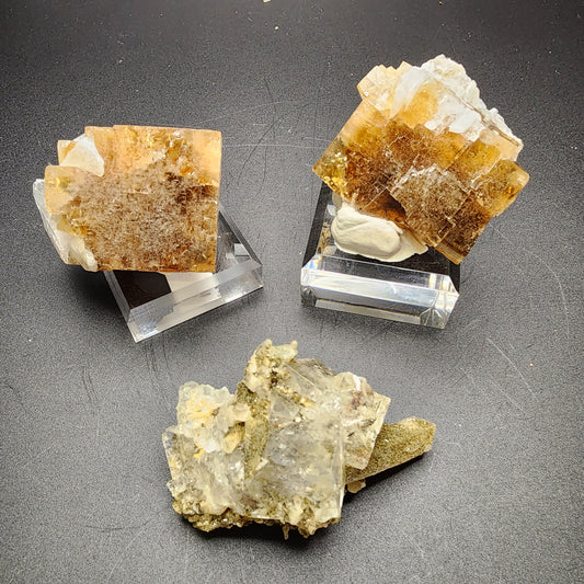 Three mineral specimens are displayed on a dark surface. The two in the back, reminiscent of the famed Clay Center Cubes, are cubic crystals with a mix of white, yellow, and brown hues, each resting on a clear stand. The specimen in the foreground is an irregular formation featuring shades of gray and white from Dalnegorsk. These remarkable pieces come from "The Crystalary" collection under their product name LIVE- sinfullilblonde- 9/5/2024.