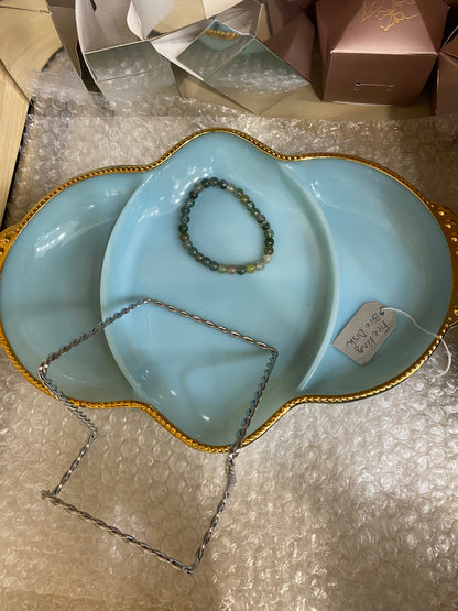 A serving dish from The Crystalary's LIVE- thetaylorkristin- 7/5/24 collection, featuring a blue three-section design with a gold rim, is placed on bubble wrap. Inside the dish, there's a Moss Agate bracelet and a wire piece shaped like a square. A small paper tag with writing is attached to the dish.