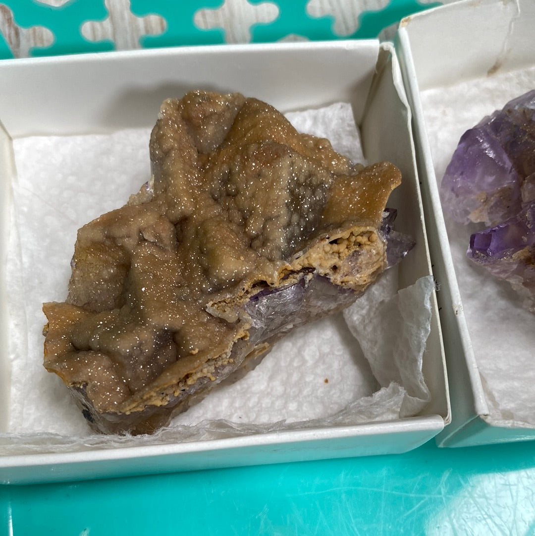 A close-up image of a brown and partially purple mineral rock displayed on white tissue paper inside a small white box reveals the rough and textured surface of the LIVE- Tracey Bachetto- 4/25/24 from The Crystalary. Another partially visible rock lies in a similar box beside it.