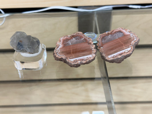 A display featuring two types of crystals on a glass shelf: on the left, a small, rough gray crystal from El Hammam on a clear stand; on the right, an agate geode revealing pink and white layers inside. This arrangement is presented by The Crystalary in their LIVE- Tracey Bachetto- 5/11/24 collection.