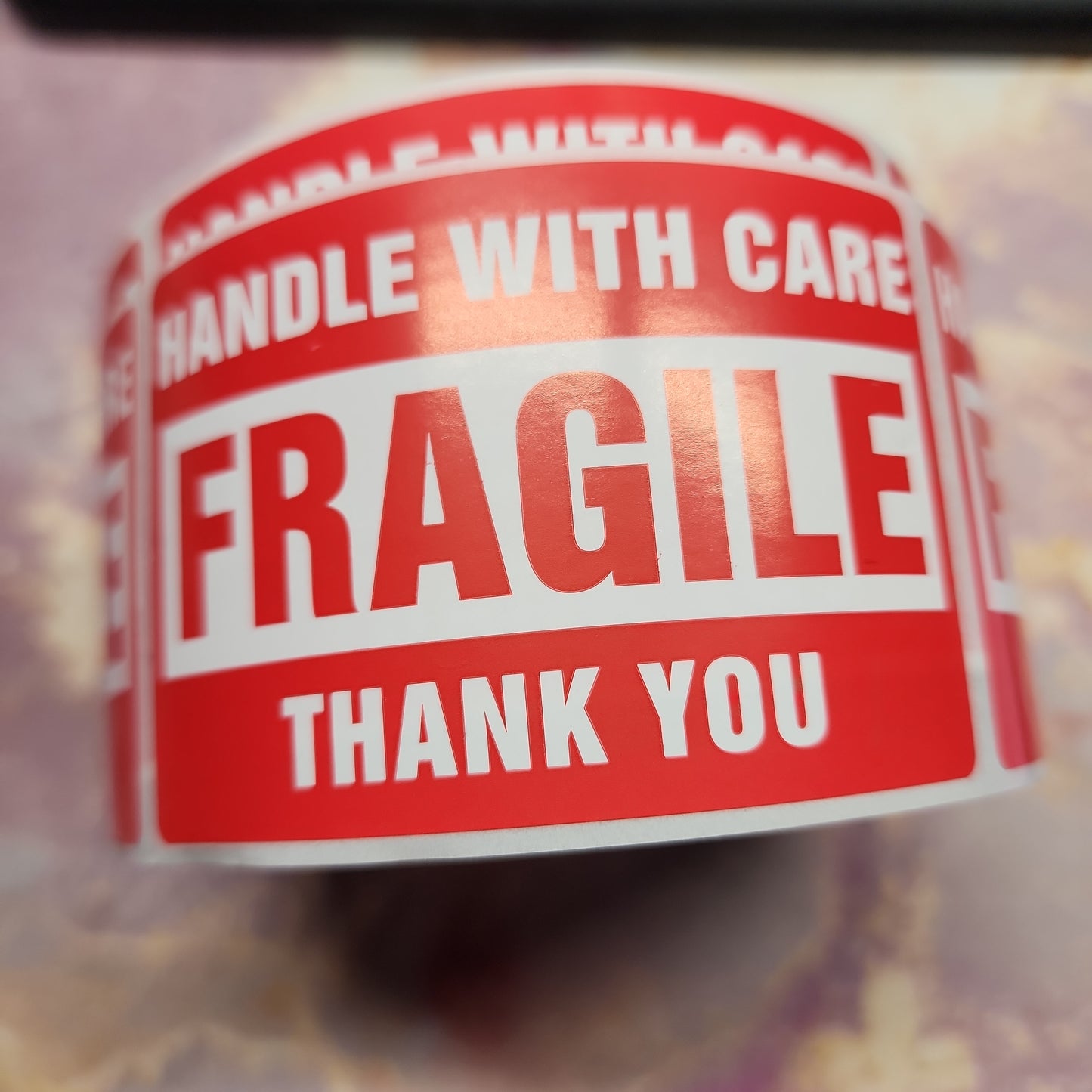 A close-up image of a roll of The Crystalary's LIVE- TracyBacchetto- 2/9/24 shipping stickers. The stickers read "HANDLE WITH CARE FRAGILE THANK YOU" in bold white letters against a red background. The roll is partially unwound, showing multiple identical labels, ready to be applied to various packages or baskets.