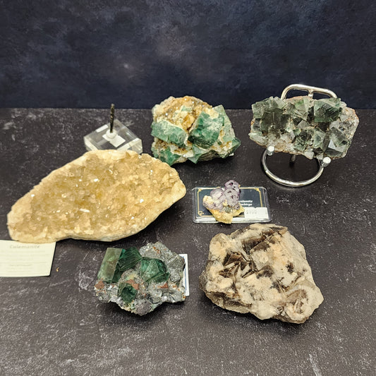 A display of mineral specimens on a dark surface, featuring five unique stones: a cluster of yellowish crystals, green cubic crystals on stone, small purple crystals, a piece with green and clear sections reminiscent of the Tourmaline Queen's realm, and a rough beige rock with brown inclusions. These stunning stones are from The Crystalary's LIVE- Vineyardguy- 6/13/24 collection.