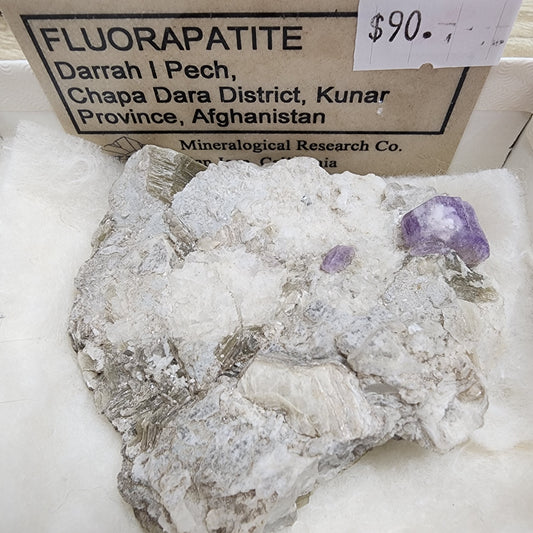 A mineral specimen labeled as "LIVE- Vineyardguy- 8/23/24" from The Crystalary, originating from Darrah I Pech, Chapa Dara District, Kunar Province, Afghanistan. Priced at $90, this exquisite fluorapatite is embedded in a white matrix adorned with visible purple crystals and is elegantly showcased in a display case.