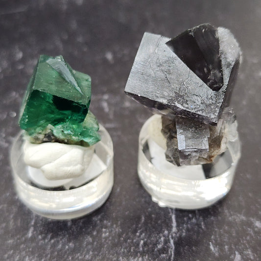 The Winzalite samples from The Crystalary, each showcased on circular stands with white putty, evoke a sense of awe. The left sample, part of the LIVE- Winzalite- 6/11/24 collection, is a green, translucent crystal reminiscent of the Northern Lights. In contrast, the right sample features dark, opaque crystals. Both samples are set against a textured, dark background for dramatic effect.
