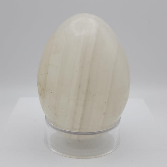 A polished Mangano Calcite Egg from The Crystalary is elegantly displayed on a small, clear stand against a plain, light gray background. This exquisite stone, sourced from Mexico, features a smooth and glossy finish with delicate vertical striations in shades of white and beige. When exposed to UV light, its fluorescent properties reveal an enchanting glow.