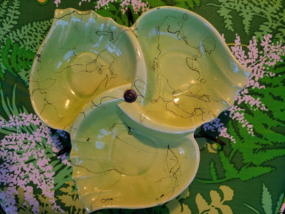 The Crystalary's Mid Century Funky Chartreuse Hollywood Ceramics Divided Serving Dish features three interconnected, leaf-shaped sections displayed on a green, leafy-patterned background. Made in California by Hollywood Ceramics, this handmade ceramic dish exemplifies the kintsugi technique with its gold-filled cracks. A small dark element connects the sections at the center, adding to its unique charm as an elegant hors d'oeuvre stand.