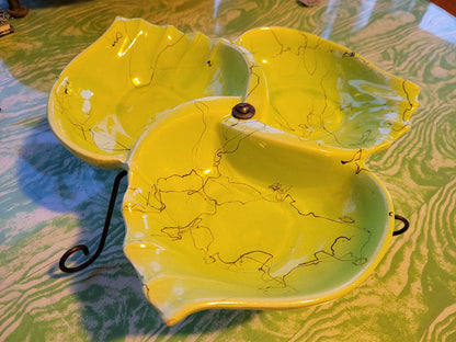 A close-up showcases the "Mid Century Funky Chartreuse Hollywood Ceramics Made in California Divided Serving Dish Hors d'oeuvre on Stand" by The Crystalary. This vibrant chartreuse ceramic dish, featuring three leaf-shaped compartments with black veining patterns, is handmade in California and elegantly displayed on a metal stand against a green marbled surface.