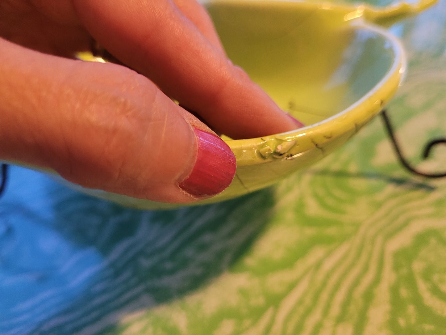 A person's hand, adorned with pink nail polish, holds The Crystalary’s Mid-Century Funky Chartreuse Divided Serving Dish made by Hollywood Ceramics in California, showing visible cracks and chips along the rim. The vibrant dish contrasts beautifully against a green and white patterned surface.