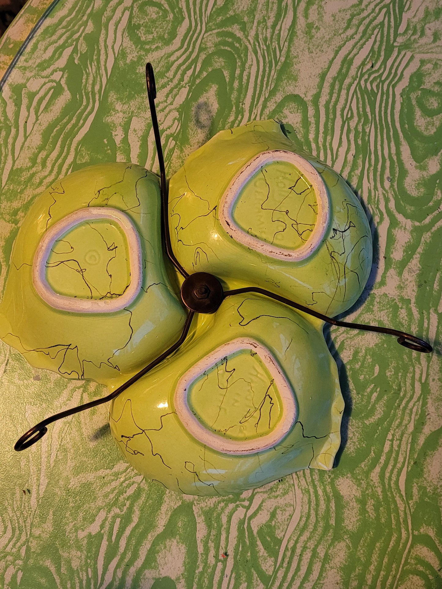 A ceiling light featuring three sockets, mounted on a textured green and white surface, seems to be damaged. The shades, which bear a resemblance to mid-century designs by The Crystalary, have yellowish hues with pink and white concentric circles. These shades are reminiscent of the "Mid century Funky Chartreuse Hollywood Ceramics Made in California Divided Serving Dish Hors d'oeuvre on stand" and show noticeable cracks and missing pieces while being held together by a central black fixture.