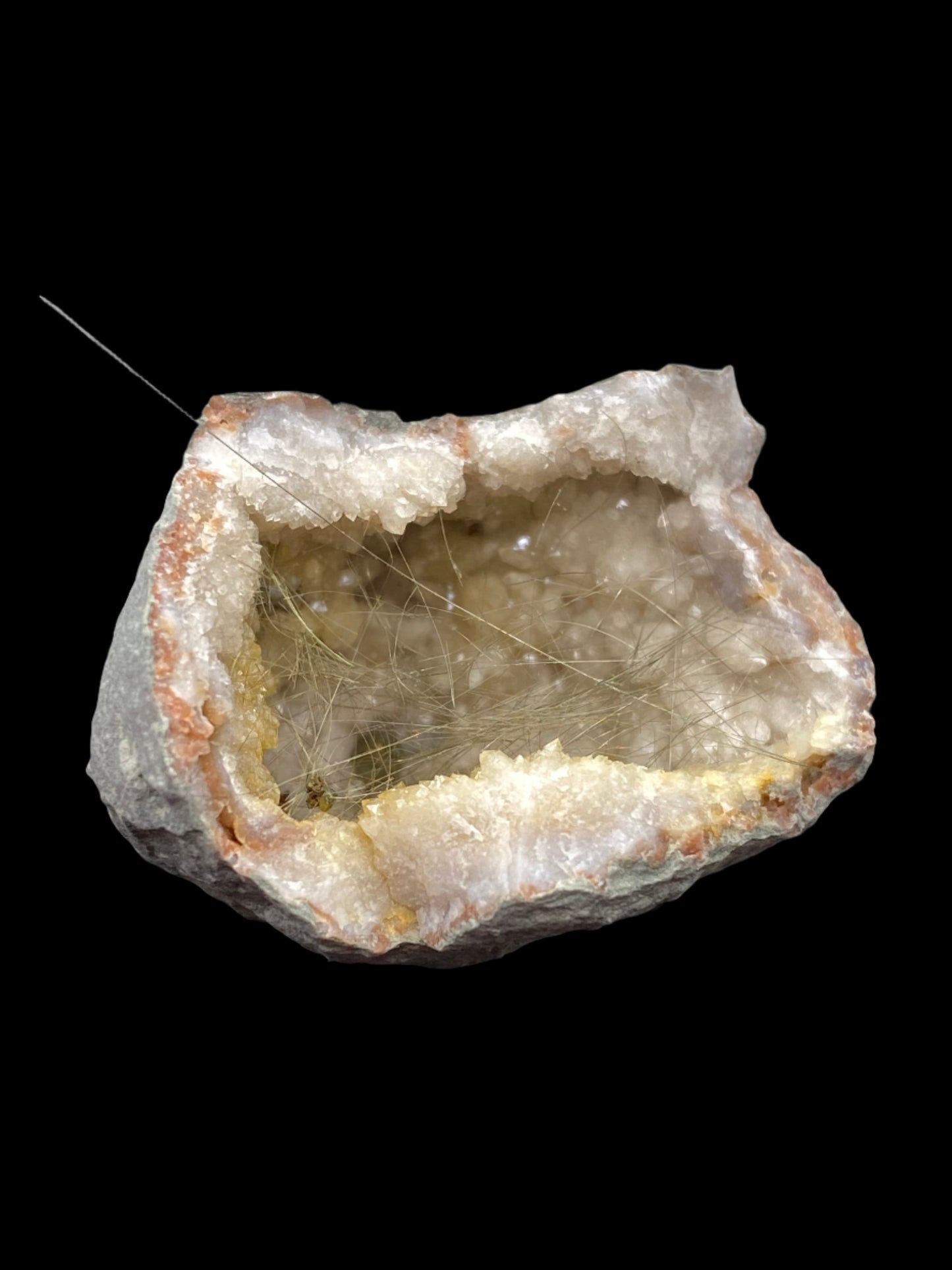 A rough geode from the collection of The Crystalary, branded as "Millerite- Halls Gap, Lincoln Co., Kentucky," showcases a grey and brown outer surface which opens to reveal an inner cavity lined with sparkling, translucent quartz crystals. Fine, hair-like inclusions give the crystals a unique texture, while the nickel-rich matrix adds to its distinctiveness. The background is black.