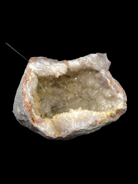 A rough geode from the collection of The Crystalary, branded as "Millerite- Halls Gap, Lincoln Co., Kentucky," showcases a grey and brown outer surface which opens to reveal an inner cavity lined with sparkling, translucent quartz crystals. Fine, hair-like inclusions give the crystals a unique texture, while the nickel-rich matrix adds to its distinctiveness. The background is black.